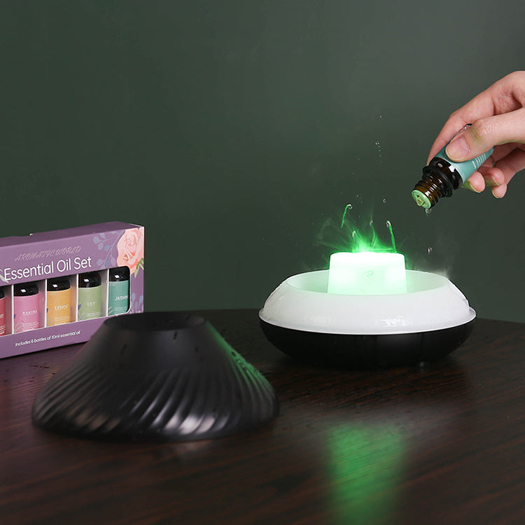 Black LavaLux Aromatherapy Diffuser, with the lid removed, on a table, turned on, with the LED light set to green. A hand is adding essential oils from a small bottle into the diffuser's reservoir. Drops can be seen splashing along with some mist, mixed with the green light. The lid is next to the diffuser. There is also a box with a set of small bottles of essential oils on the table.