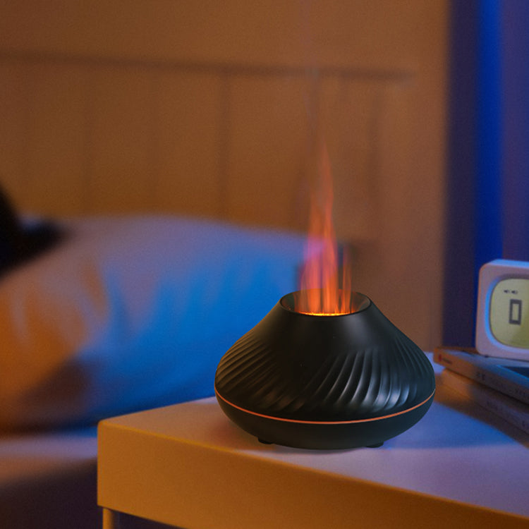 Black LavaLux Aromatherapy Diffuser on a table in a bedroom, turned on, with the LED light set to orange, and mist coming out like a lit flame. In the background, the bed is visible and blurred, and on the table next to the diffuser are two books and a digital alarm clock.